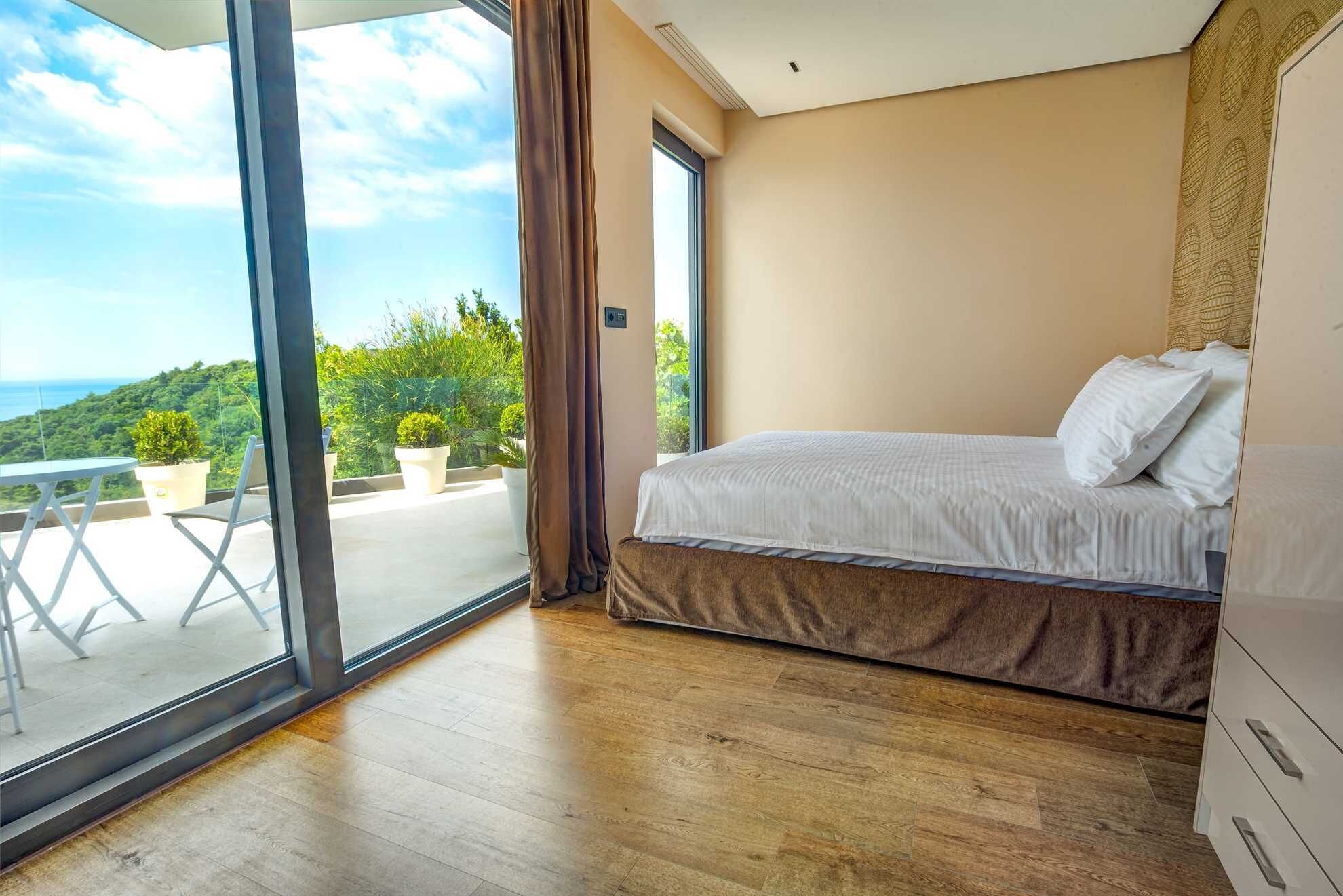 A bedroom with a large sliding glass door leading to a balcony with potted plants and a view of greenery.