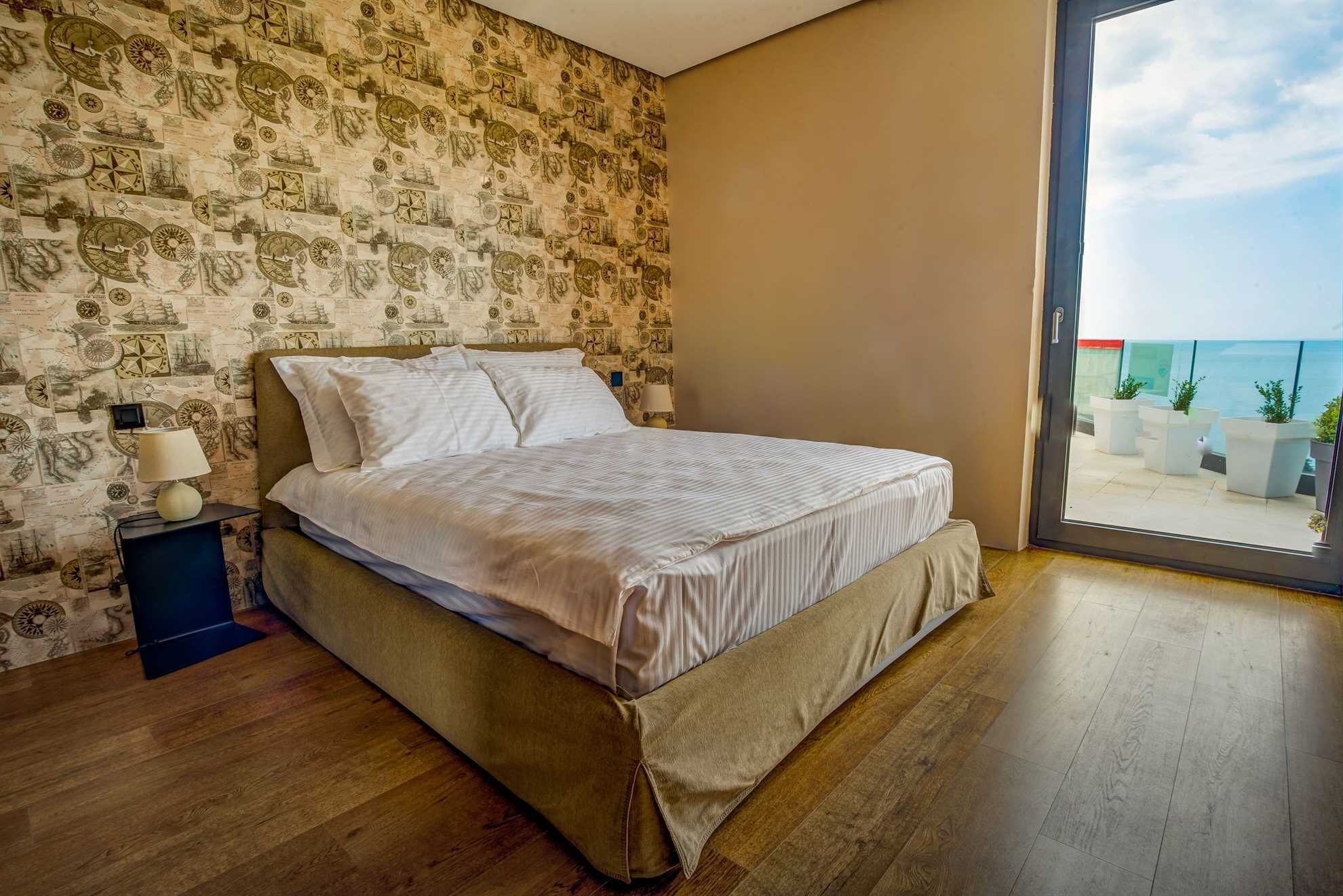 Een slaapkamer met een bed, nachtkastje, muur met zeewaas behang en uitzicht op zee door een glazen deur.