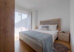 A bedroom with a wooden headboard, a bed with a grey blanket, a nightstand with a lamp, and a window with sheer curtains.