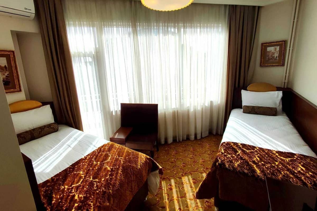 Camera d'albergo con due letti, una scrivania, una sedia, un'ampia finestra con tende e una lampada a sospensione.