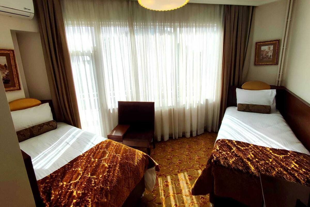 Camera d'albergo con due letti, una scrivania e una sedia. Grande finestra con tende, condizionatore d'aria e un quadro appeso al muro.