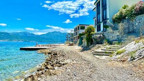 Un bâtiment situé sur la côte, avec une plage rocheuse, un ciel bleu dégagé et des montagnes en arrière-plan.