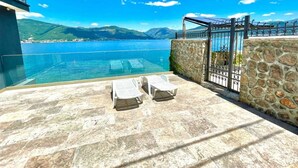 Une terrasse dotée d’un mur en pierre et d’une balustrade en verre, offrant une vue imprenable sur une mer d’un bleu profond et sur des montagnes lointaines.