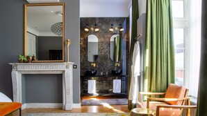 A modern bathroom with a large mirror, two sinks, and a bathtub. There is a fireplace, a window with curtains, and a seating area with a chair and a small table. - The Old Beech (Antwerp)