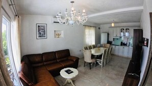 Uma sala de estar com sofá de couro marrom, uma mesa de jantar com cadeiras, um candelabro e uma área de cozinha com armários e eletrodomésticos.