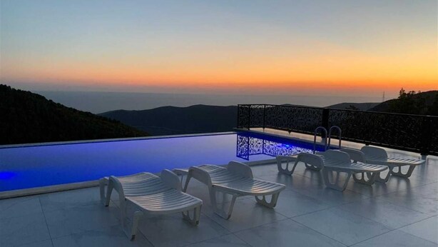 Piscina infinita no terraço com cadeiras de sol brancas e vista para o pôr do sol sobre as montanhas.