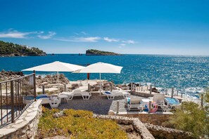 A seaside terrace with white umbrellas, lounge chairs, and a pool area.