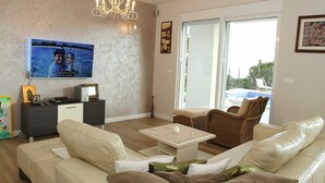 Een woonkamer met een bank, een salontafel, een televisie, een kroonluchter en uitzicht op een zwembad door een glazen deur.