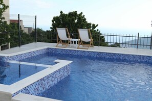 Una piscina con mosaicos azules, dos sillas reclinables blancas y vista al mar.