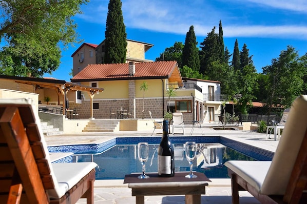 A poolside area with lounge chairs, a table with a bottle and glasses, and a view of a building with a red-tiled roof.