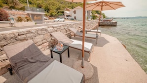 A seaside terrace with lounge chairs and umbrellas, a stone wall, and a view of the sea and buildings in the background. - Villa Stella Adriatica (Tivat)