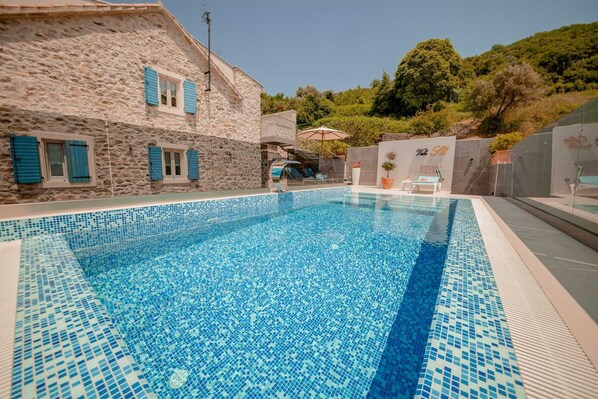 Une piscine aux carreaux de mosaïque, un bâtiment en pierre avec des volets bleus, et une vue sur une colline.