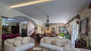 Een ruime woonkamer met een hoge plafond, stenen muren en een trap die naar een ander niveau leidt. Er zijn twee banken, een kroonluchter en een eethoek met een tafel en stoelen.