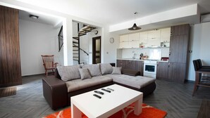 Una sala de estar moderna con un sofá modular, una mesa de centro blanca y una alfombra roja. Una escalera conduce a una cocina abierta con gabinetes de madera y una estufa.