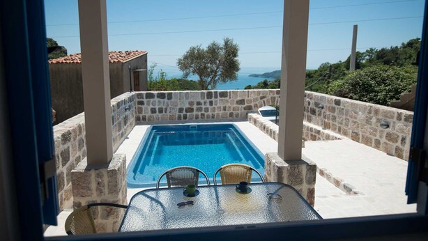 Una piscina de piedra con vistas al mar, rodeada de muros de piedra y un porche cubierto con mesa y sillas.