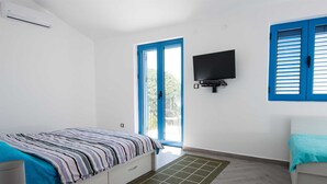 Un dormitorio con cama, televisor montado en la pared, ventana con contraventanas azules y puerta que da al exterior.