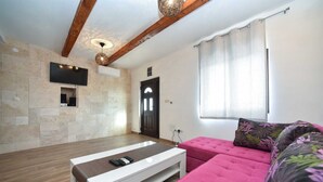Una sala de estar moderna con pared de piedra, vigas de madera, televisor, sofá rosa y ventana con cortinas.