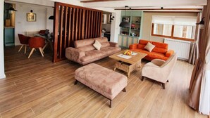 Een ruime woonkamer met houten plafond, een hoekbank, een salontafel en een eethoek met een rode stoel.
