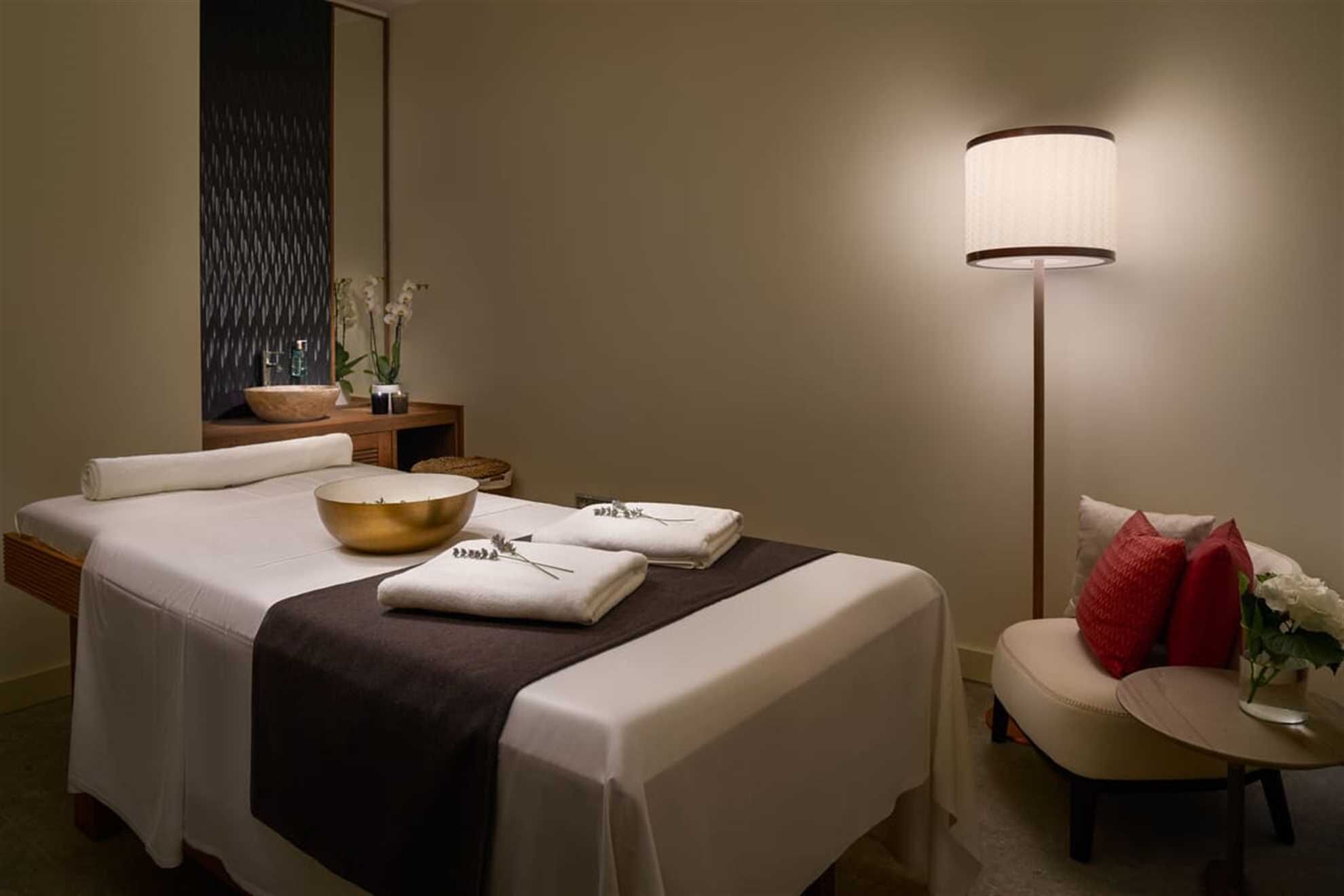 A hotel room with a neatly made bed, a bedside table with a bowl and a vase of flowers, a chair with a red pillow, and a floor lamp.
