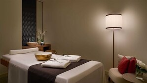 Une chambre d’hôtel avec un lit soigneusement fait, une table de chevet sur laquelle se trouvent un bol et un vase de fleurs, une chaise avec un coussin rouge et une lampe sur pied.