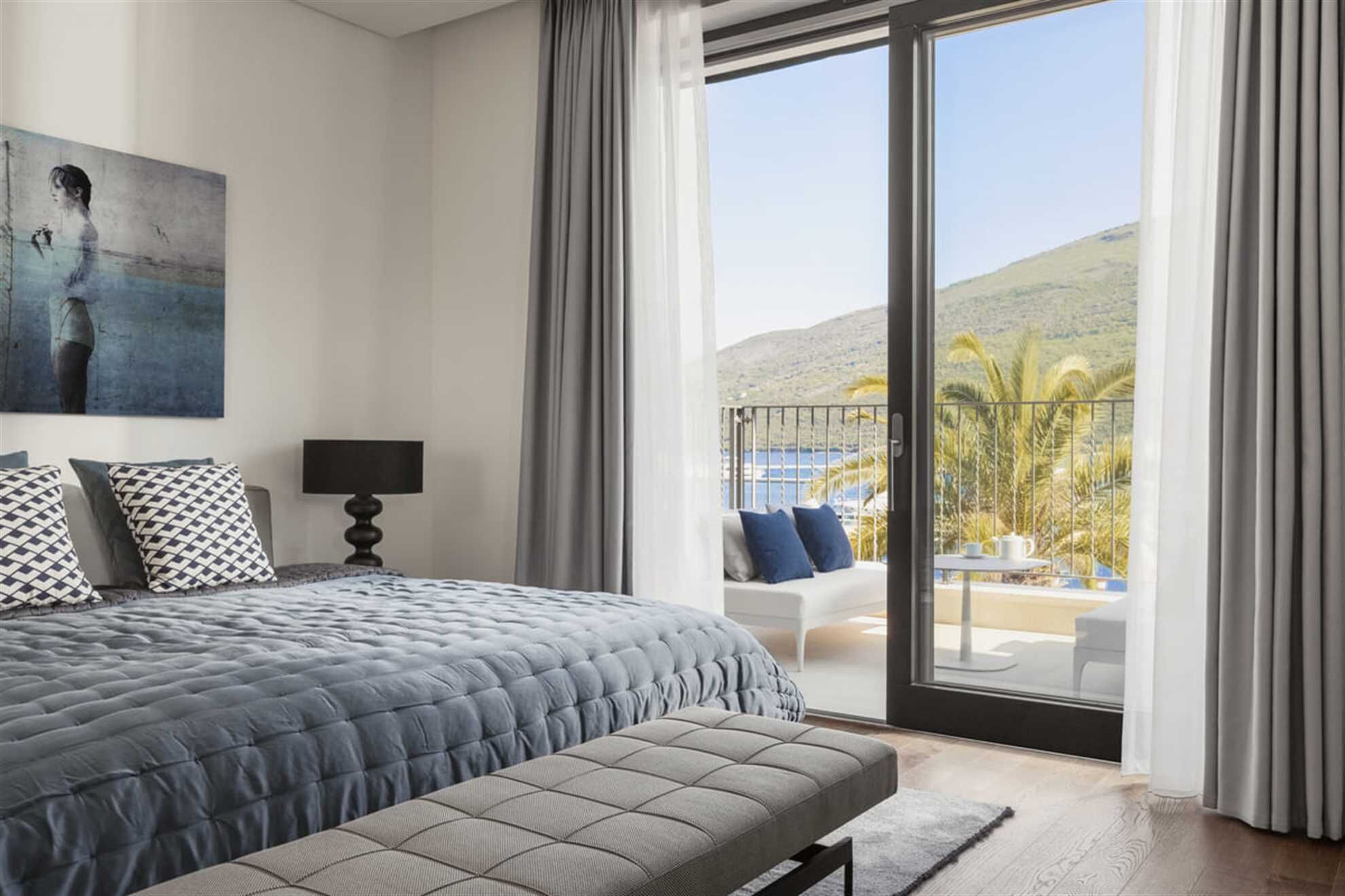 Een slaapkamer met een groot bed, een nachtkastje met een lamp, een schilderij aan de muur en een balkon met tuinmeubilair.