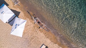 Vue aérienne d’une plage aux eaux claires et peu profondes, avec quelques parasols et une petite construction située près du bord de l’eau.