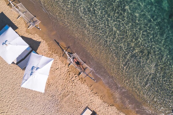 Vue aérienne d’une plage aux eaux claires et peu profondes, avec quelques parasols et une petite construction située près du bord de l’eau.
