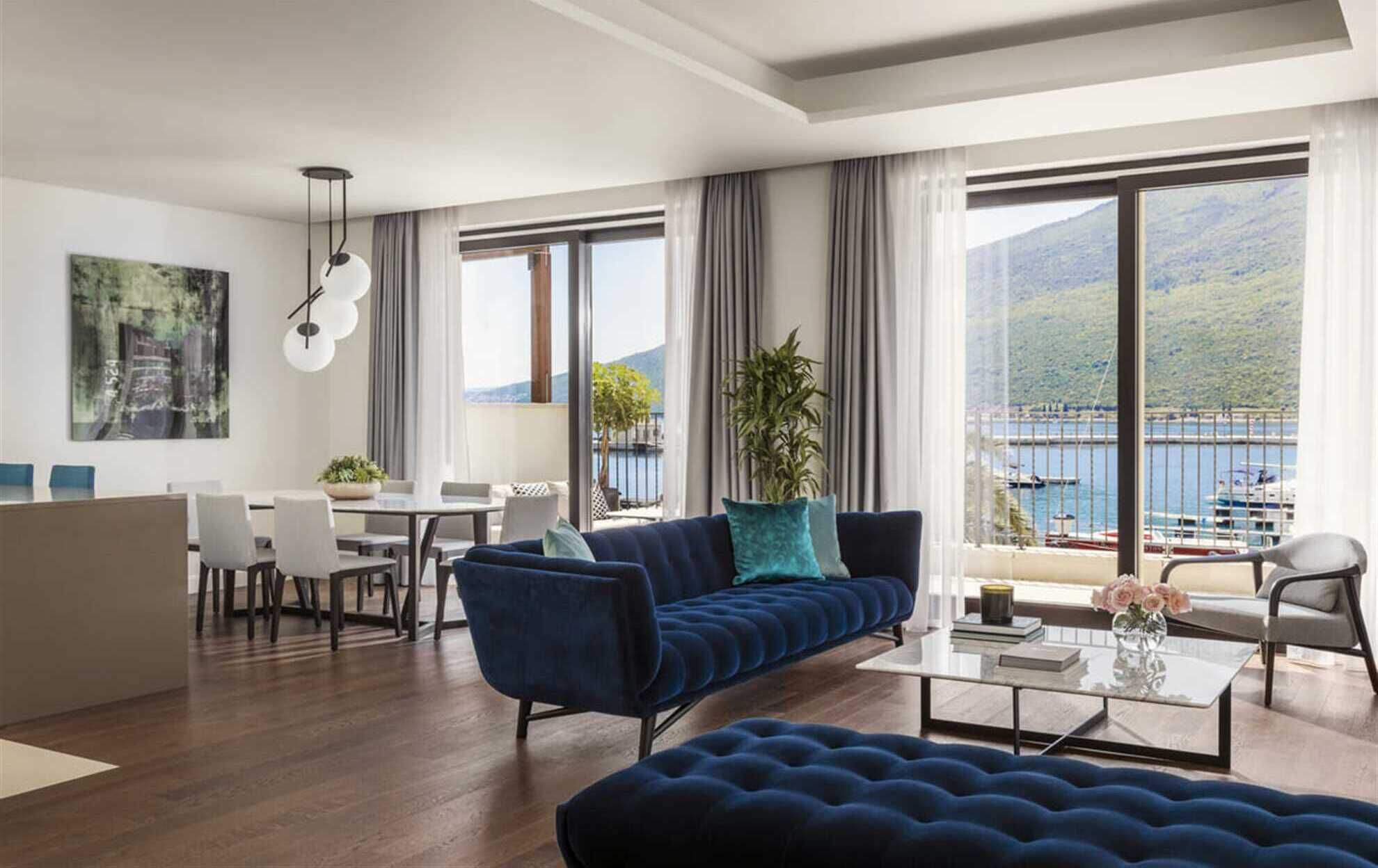 Ein modernes Wohnzimmer mit einem blauen Sofa, einem Essbereich mit weißen Stühlen und großen Fenstern, die Blick auf ein Gewässer und Berge bieten.
