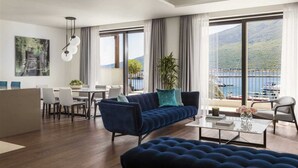 Un salon moderne avec un canapé bleu, un coin repas avec des chaises blanches et de grandes fenêtres offrant une vue sur un plan d’eau et des montagnes.