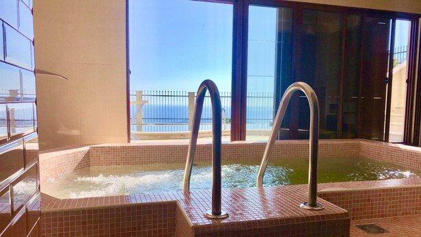 Un jacuzzi con dos manijas metálicas, con azulejos en un diseño de tablero de ajedrez, en una habitación con ventanas amplias que dan a un balcón con barandilla.