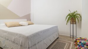 Een net opgemaakt bed met een witte deken en twee kussens, een pot met een varen en een kleurrijke bol wol op de vloer.
