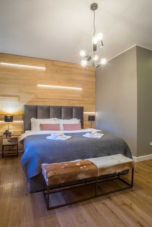 Un dormitorio moderno con cabecera de madera, una cama grande con colcha gris, un banco con manta de piel y una lámpara de techo original.