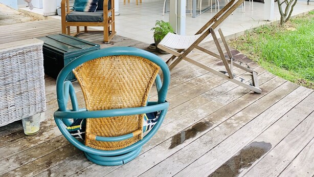 Een overdekt terras met rieten stoelen en een tafel, een houten terras en een uitzicht op het interieur van het huis.