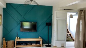 Een woonkamer met een wand in teal, een flatscreen televisie, een houten console tafel, een geweven bankje en een moderne hanglamp.