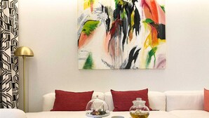 Una sala de estar moderna con un sofá blanco, cojines rojos, una mesa de centro con una revista, un hervidor y una taza. Un cuadro abstracto y colorido cuelga en la pared.