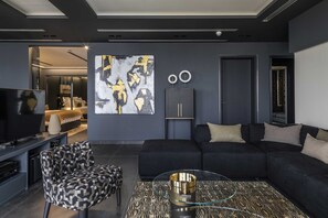 Ein modernes Wohnzimmer mit einem dunkelgrauen Sofa, einem gemusterten Sessel, einem Glastisch und einem großen abstrakten Gemälde an der Wand.