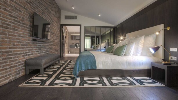 Une chambre à coucher moderne avec un mur en briques, un grand lit avec de nombreux coussins, un banc et un tapis à motifs.