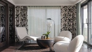 Un salon moderne avec une table basse ronde, deux fauteuils blancs, un lampadaire et une grande fenêtre agrémentée de rideaux à motifs floraux.