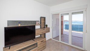 A modern living room with a flat-screen TV, wooden furniture, a small plant, and a view of the sea through a glass door. - Beautiful apartment in Marina (Marina)