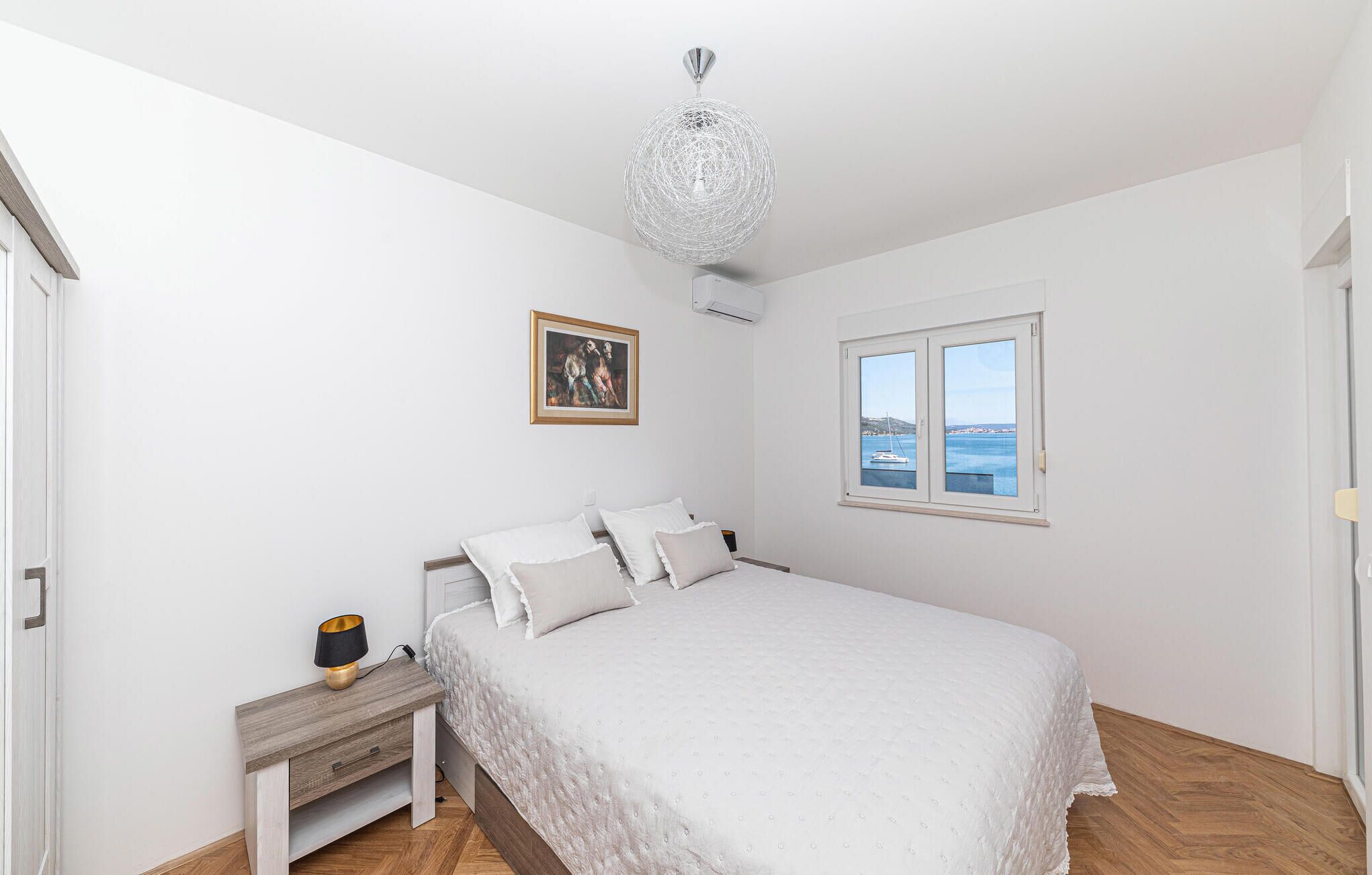 Ein Schlafzimmer mit einem Bett, einem Nachttisch, einer Lampe, einem gerahmten Bild, einem Fenster mit Meerblick und einer Pendelleuchte.
