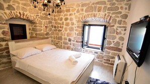 Un dormitorio con pared de piedra, cama con ropa de cama blanca, una araña, un televisor y una ventana.