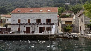 Un edificio de piedra con techo de tejas, un muelle que se adentra en el agua y montañas al fondo.
