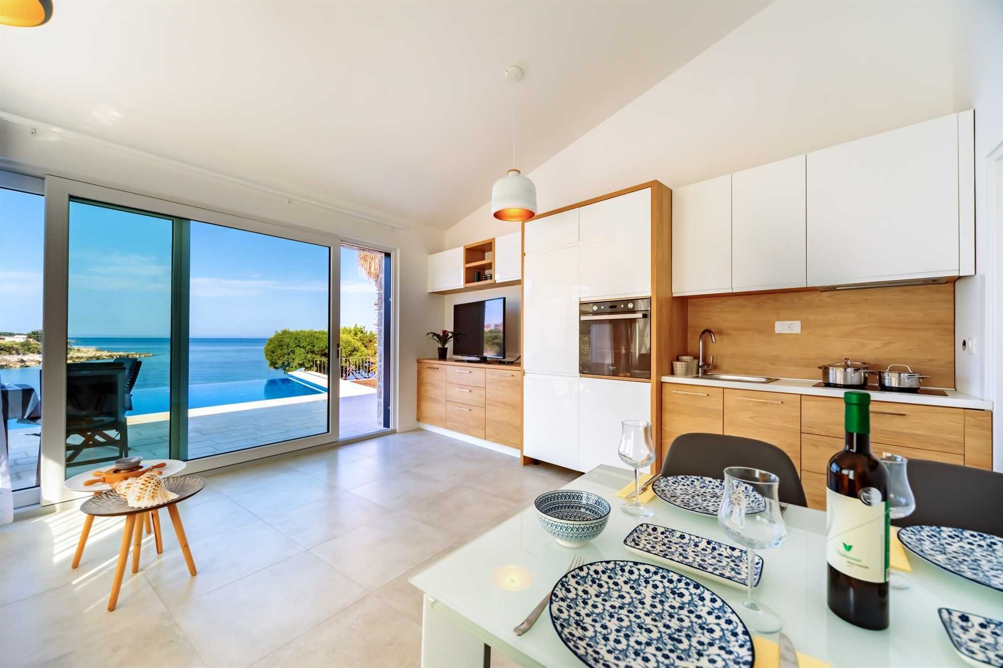 A modern kitchen with a view of the sea, a dining area with a table set for two, and a large sliding glass door leading to an outdoor seating area.