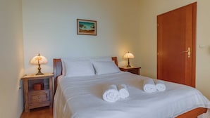 Een net opgemaakt bed met witte lakens en twee opgerolde handdoeken. Er zijn twee nachtkastjes met lampen en een klein houten nachtkastje met een lamp. Een deur is rechts zichtbaar.