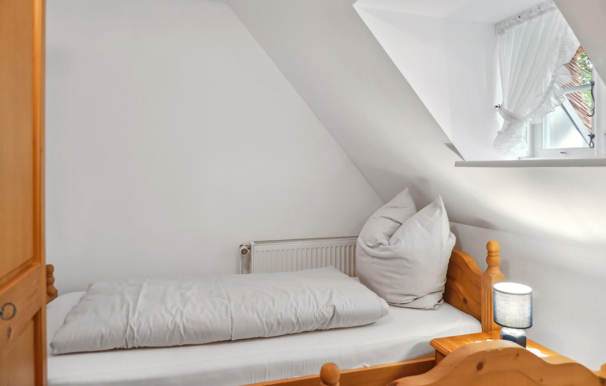 Une petite chambre avec un lit simple, un plafond en pente, une fenêtre avec des rideaux légers, une tête de lit en bois et une table de chevet avec une lampe.