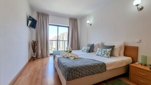 A modern bedroom with a large bed, a bedside table, a nightstand, a TV mounted on the wall, and a window with curtains.