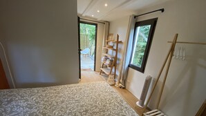 A bedroom with a patterned bedspread, a wooden ladder, a shelf with towels, and a view of the outdoors through a window. - Mini Villa With Jacuzzi (Cagnes sur mer)