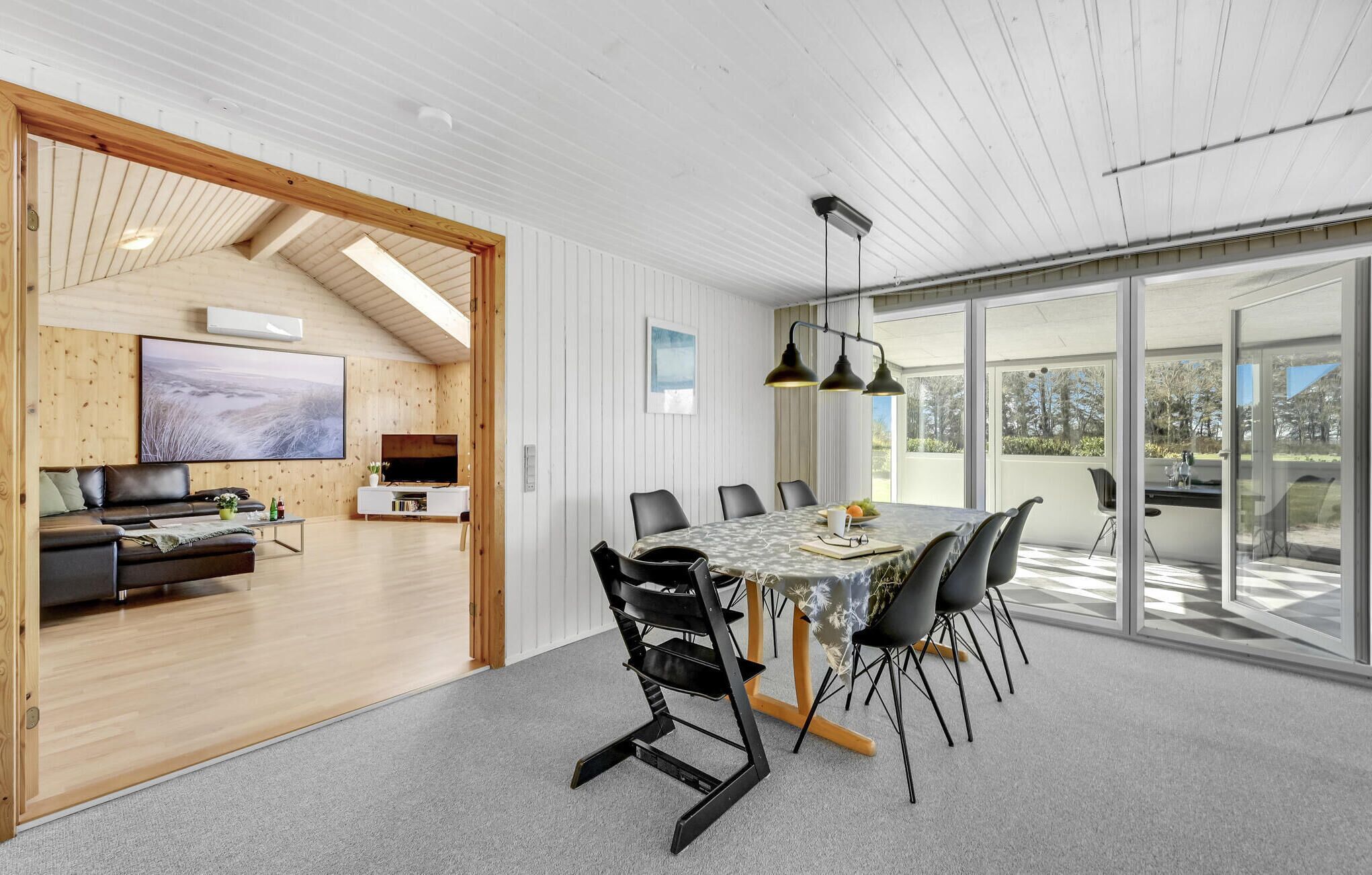 A modern interior with a dining area, a living room with a TV, and large windows offering a view of the outdoors.