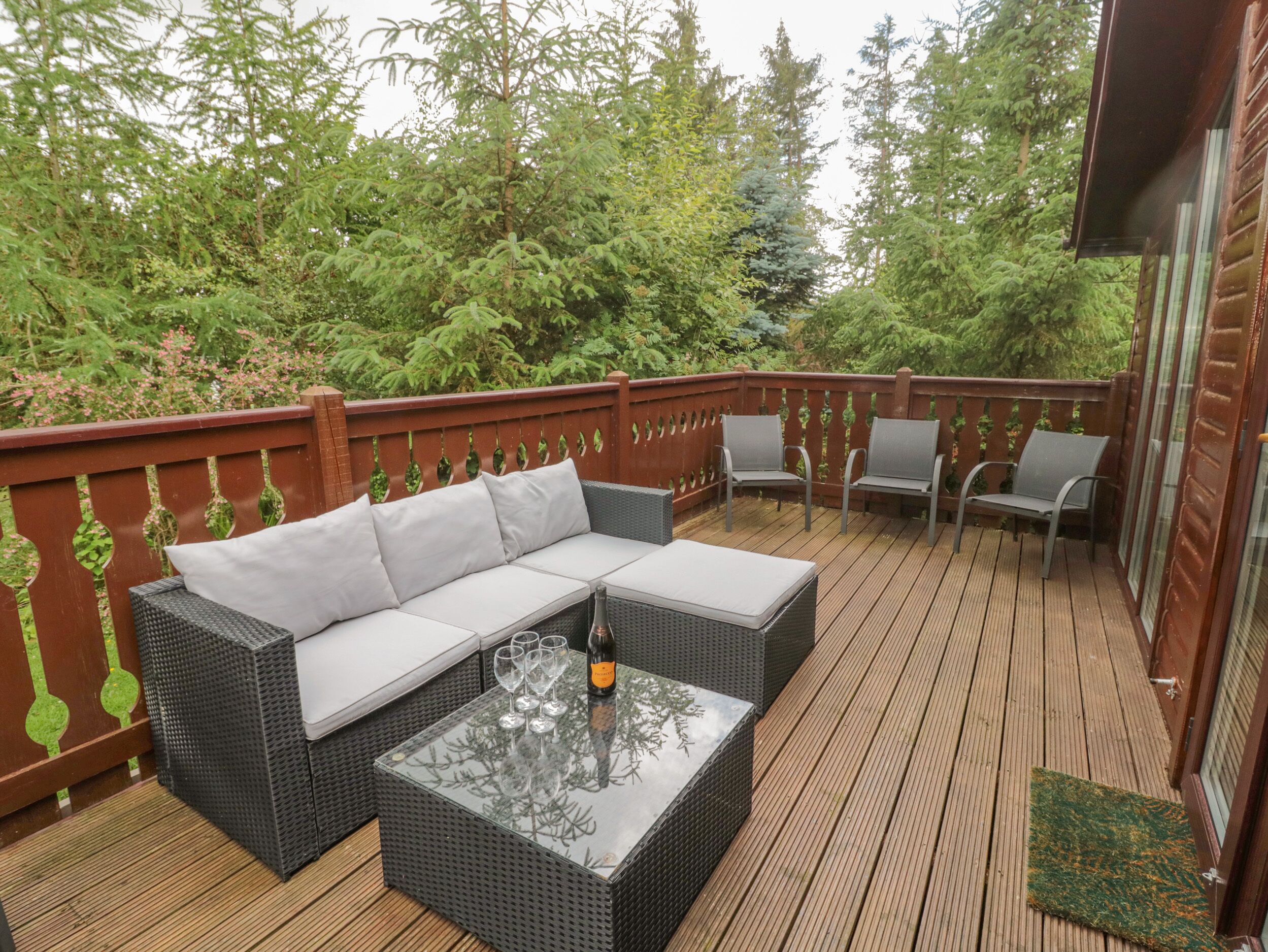 A covered deck with a wicker sofa, two chairs, and a glass-top table with wine glasses and a bottle.
