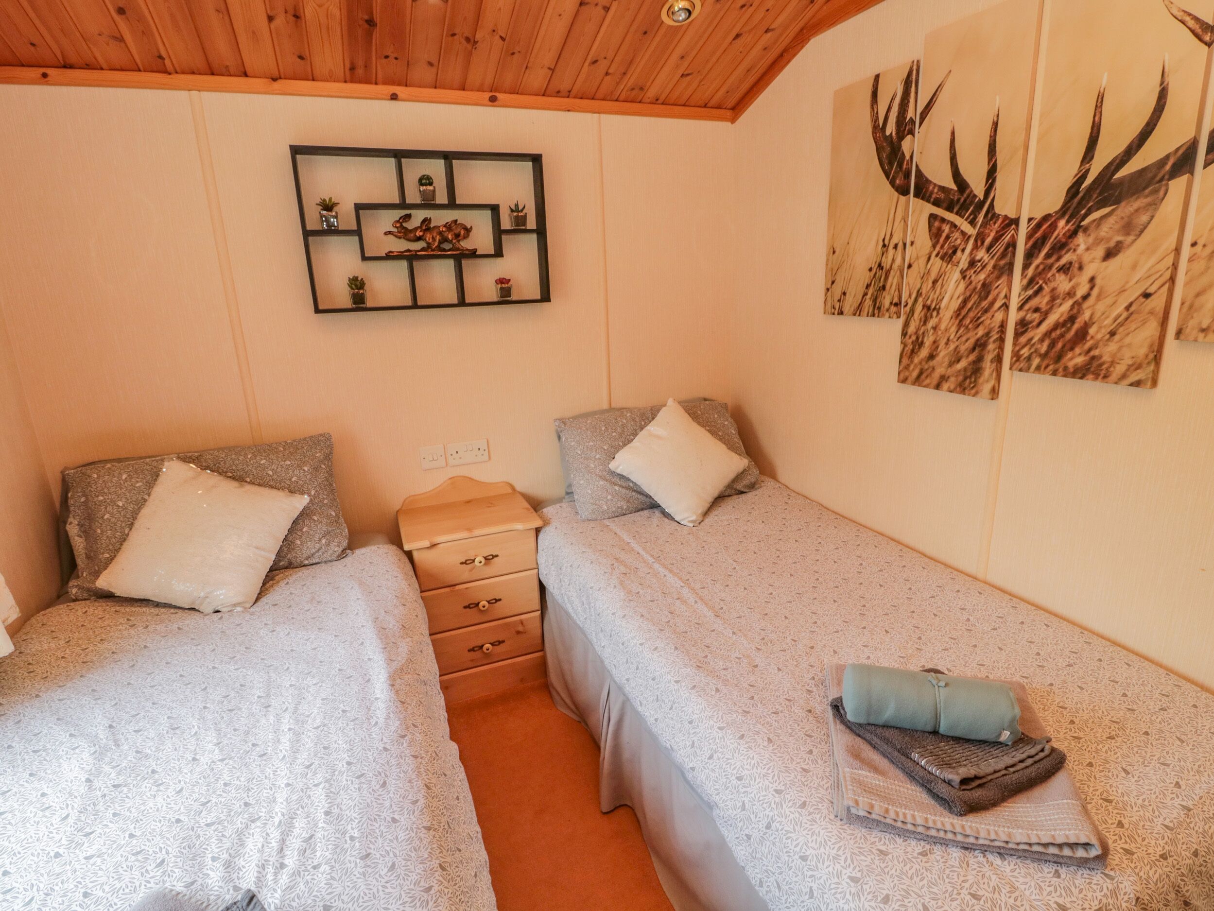 A cozy bedroom with two beds, a wooden ceiling, a shelf with decorative items, and a wall painting.